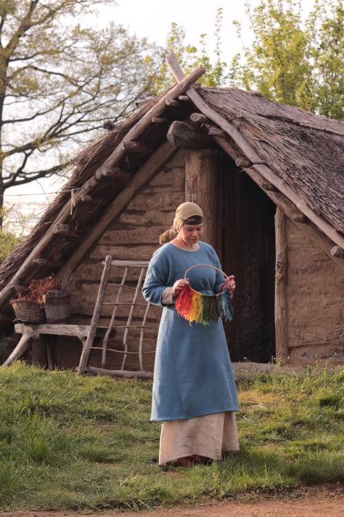 Kundenfoto: Eine Frau steht vor einem frühmittelalterlichen Haus. Sie trägt ein knöchellanges Unterkleid aus Leinen, genäht von Hannyrdi. Das blaue Überkleid ist nicht aus meiner Näherei.